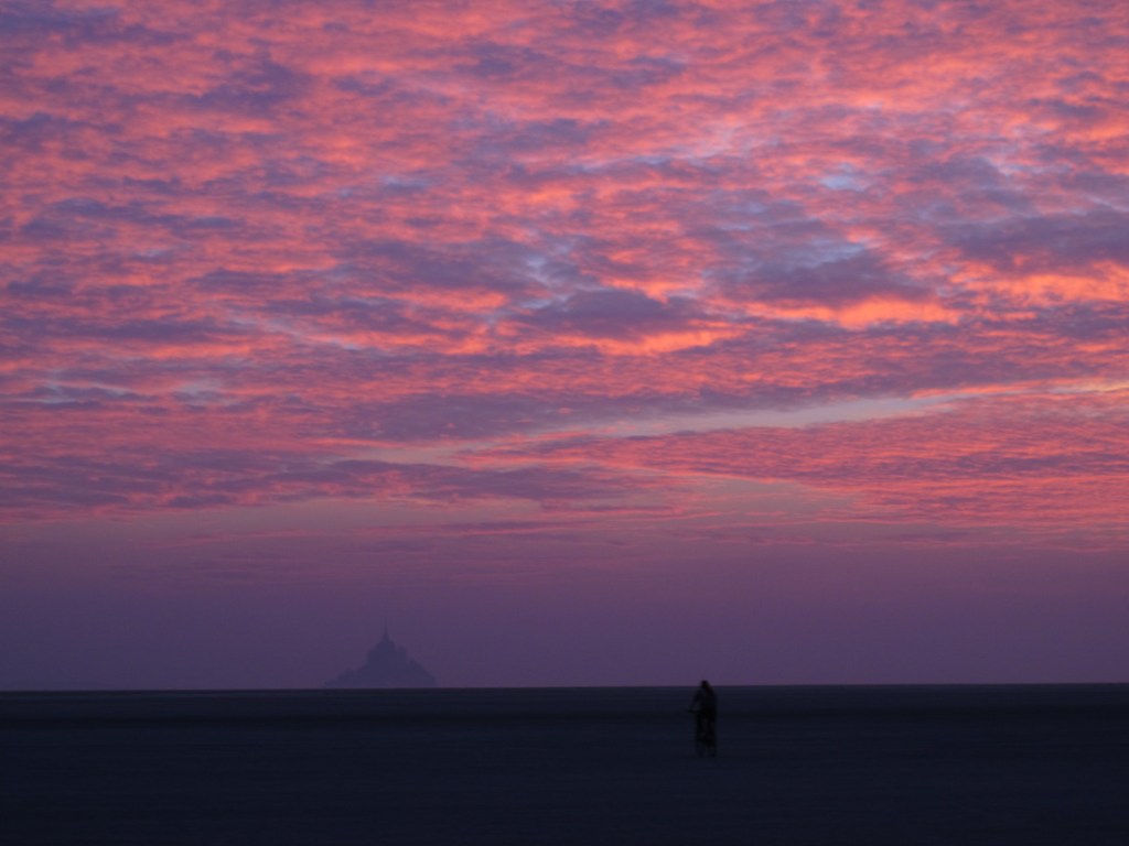 Sunset behind Le Mont-Saint-Michel, France, with lone cyclist on the mud flats.